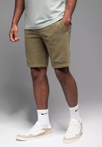 Shorts kaki en matériau doux avec des coutures en angle et un petit logo. Associés à des chaussettes de sport blanches et des baskets beiges.