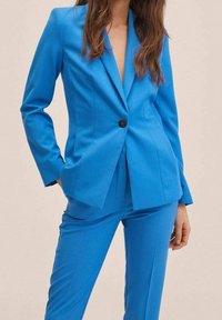Blazer bleu cintré et pantalon assorti en tissu lisse. Le blazer est doté d'un bouton noir unique et d'une coupe ajustée.