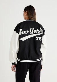 Veste de varsity noir et blanc avec "New York" en lettres blanches texturées, design sportif, poignets et ourlet côtelés. Le modèle porte un pantalon noir.