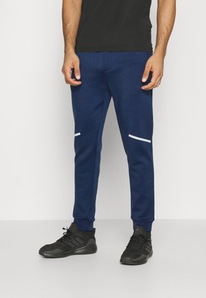 Homme portant un pantalon de jogging bleu marine avec des bandes réfléchissantes blanches et des chaussures de sport noires, debout devant un fond clair uni.