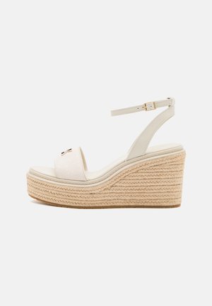 Beige kilehælsandal med flettet platformsål, ankelrem med guldbeklædt spænde og åben tå med en lille metal sløjfedetalje.