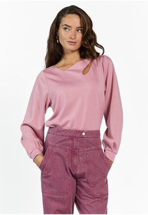 Vrouw met lang golvend haar, gekleed in een roze top met lange mouwen en een keyhole-uitgesneden hals, en een hooggesneden mauve broek, handen in de zakken.