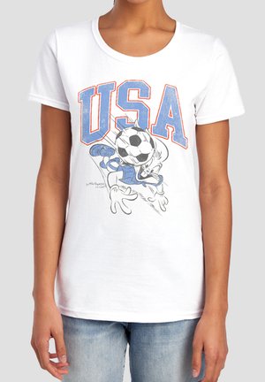 Weißes T-Shirt mit großem blauem und rotem "USA"-Text und einer Cartoonfigur, die einen schwarz-weißen Fußball köpft, getragen mit hellblauen Jeans.