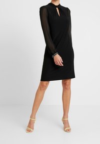 Frau in einem knielangen schwarzen Kleid mit durchsichtigen langen Ärmeln, Ausschnitt mit Schlitz und hellgelben Riemchen-High Heels, stehend vor weißem Hintergrund.
