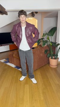 Βουργουνδίας δερμάτινο μπουφάν με ροζ λεπτομέρειες, λευκό μπλουζάκι, γκρι τζιν και λευκά αθλητικά, στέκεται σε ξύλινο πάτωμα σε ένα δωμάτιο με φυτά.