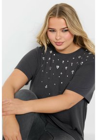 Grå t-shirt med kort ärm tillverkad av mjukt material, prydd med metalliska detaljer utspridda över framsidan, med rund halsringning.