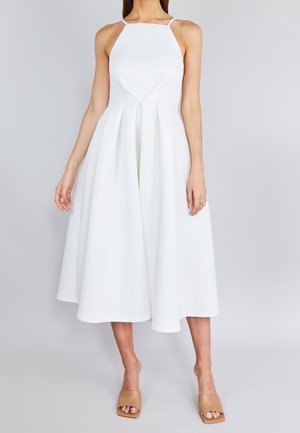 Frau trägt ein ärmelloses weißes Midikleid mit figurbetontem Oberteil und ausgestelltem Rock, kombiniert mit beigen offenen Sandalen.