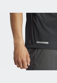 Schwarzes Sportshirt mit glatter Textur, kurzen Ärmeln, seitlichem Schlitz und einem Etikett mit "WIND.RDY" am Saum. Hand ruht auf dem Shirt.
