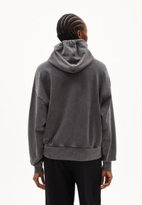 Person mit geflochtenem Haar, der einen lockeren, dunkelgrauen Hoodie und schwarze Hose trägt, von hinten gegen einen weißen Hintergrund gesehen.