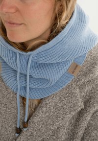 Lichtblauwe, gebreide infinity-sjaal met textuurpatronen en verstelbare trekkoorden. Bevestigd label in een neutrale kleur. G gedragen over een grijze, donzige trui.