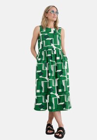 Grünes ärmelloses Kleid mit einem weißen geometrischen Muster, knielang, mit einem gerafften Rock und seitlichen Taschen. Das Model trägt schwarze Sandalen.