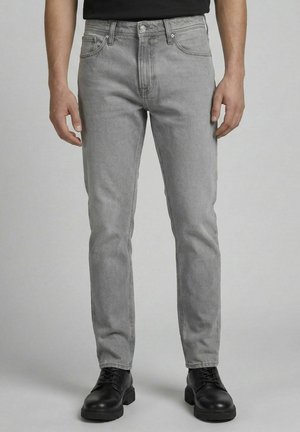 Homme portant un jean slim gris clair et des bottes en cuir noires à lacets, debout devant un fond uni.