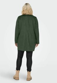 Chaqueta larga de ante verde con corte recto, llevada sobre leggings negros y botines de color canela, mostrando una textura suave y detalles de diseño mínimos.