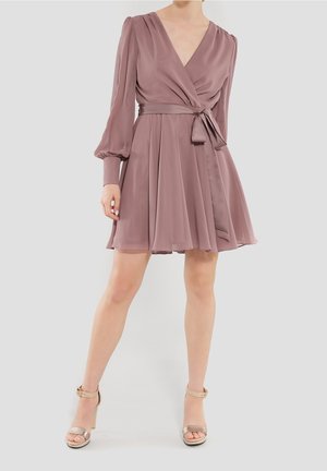 Kobieta ubrana w wrzosową sukienkę kopertową z długim rękawem i wiązaną w talii szarfą oraz beżowe sandały na wysokim obcasie na jednolitym tle.