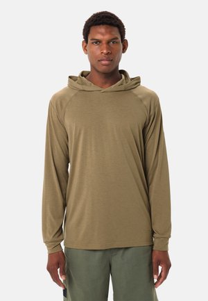 Uomo che indossa una maglia con cappuccio a maniche lunghe verde oliva e pantaloni verdi, in piedi di fronte a uno sfondo semplice.