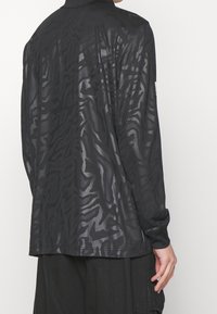 Homme portant une chemise noire à manches longues avec des motifs abstraits sombres subtils, associée à un pantalon noir ample, vu de dos.