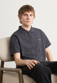 Tmavě šedé polo tričko s límečkem na knoflíky a krátkými rukávy, s malým logem na levé hrudi. Nosené v sedě na židli.