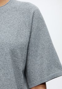Gros plan d'une personne portant un pull en maille gris chiné à manches courtes avec un col rond, sur un fond uni.