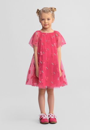 Jeune fille debout, face à l'avant, portant une robe rose à fleurs avec des manches en tulle et des baskets roses, sur un fond clair uni.