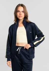 Mujer joven con cabello castaño claro que lleva un chándal azul marino con rayas verdes y crema, y un top corto crema, sobre un fondo gris liso.