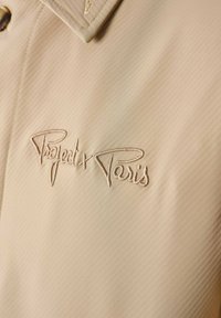 Chaqueta beige de tejido texturizado, con un cuello distintivo y el logo bordado "Project x Paris" en un color a juego.