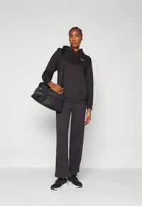 Felpa nera con cappuccio e pantaloni abbinati in tessuto morbido, abbinati a una borsa nera. L'outfit include scarpe sportive nere con dettagli bianchi.