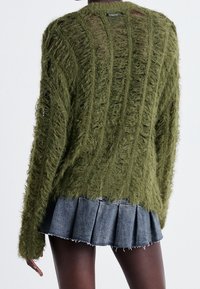 Grüner, strukturierter Strickpullover mit offener Maschenstruktur und rohen Kanten, kombiniert mit einem Jeans-Faltenrock.
