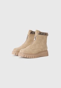 Tan ankle boots made of suede, featuring metallic side zippers and a textured knit collar, set on a chunky, ridged sole.