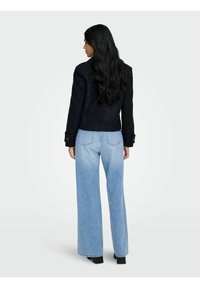 Veste texturée marine avec un design à double boutonnage, associée à un jean large bleu clair. Accent sur les boutons visibles et poches arrière sur le jean.