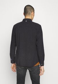 Mörkblå långärmad skjorta med en prickig textur, med krage, knappmanschetter och en rundad nederkant. Bakifrån syns axelsömmar och logotag.