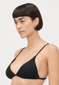 Black triangular bikini top with ribbed texture, thin adjustable straps, and a small gold logo accent on the left side.