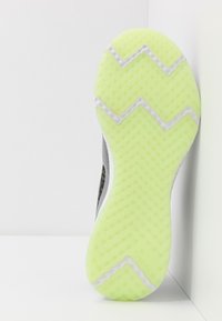 Semelle d'une chaussure de course avec une semelle extérieure texturée jaune-vert vif et des motifs en zigzag blancs, positionnée debout contre un coin blanc.
