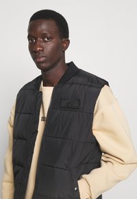 Svart pufferväst med dragkedja fram, med en hexagonal patch och ribbad krage, bärs över en ljusbeige sweatshirt med enkel textur.