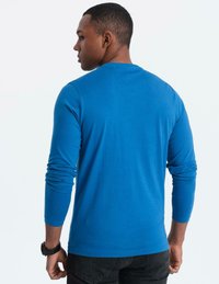 Man som står sidan med en långärmad blå skjorta, svarta jeans och en svart klocka på vänster handled.