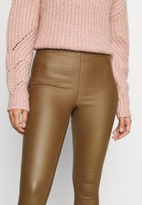 Leggings in finta pelle marrone con un design aderente e slanciato. Abbinati a un maglione lavorato a maglia rosa chiaro con motivi intricati sulle maniche.