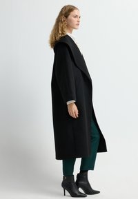 Zwarte, lange jas met brede revers en zakken, gecombineerd met donkergroene broek en zwarte enkellaarsjes met hak. Gladde textuur overal.