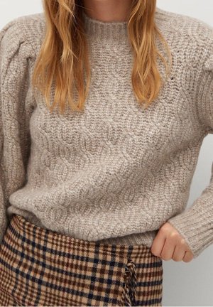 Žena v béžovém svetr s kabelovým vzorem a hnědé kostkované sukni pózuje s jednou rukou na bocích před neutrálním pozadím.