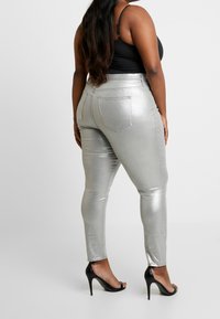 Jeans ajustados metálicos plateados con una cintura alta, que cuentan con bolsillos traseros, una textura suave y un corte ceñido, combinados con sandalias de tacón negras.