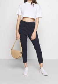 Mörkblå cropped byxor med dragsko i midjan, tillsammans med en vit kortärmad topp och vita sneakers. En beige texturerad handväska fulländar looken.