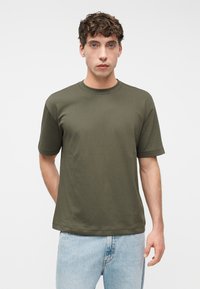 Νέος άντρας με καστανά σγουρά μαλλιά, που φοράει μπλουζάκι ελιάς με κοντά μανίκια και ανοιχτό μπλε τζιν, στέκεται μπροστά σε απλό λευκό φόντο.