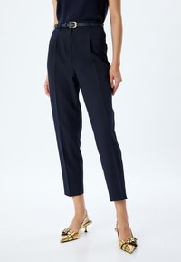 Pantalones de corte navy con una cintura alta, pliegue marcado y un cinturón negro con hebilla dorada; combinados con tacones de slingback dorados.