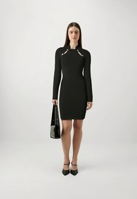 Abito nero aderente a manica lunga con ritagli sulle spalle, impreziosito da accenti metallici. Modella che tiene una piccola borsa nera, indossando décolleté nere.