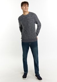 Tamsiai pilkas mezginys su tekstūrine raštais, derinamas su mėlynomis džinsomis ir tamsiai mėlynais sportiniais bateliais. Modelis stovi su rankomis kišenėse, šypsodamasis.