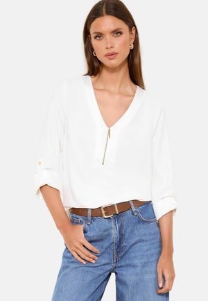 Kobieta ubrana w białą bluzkę z dekoltem w serek i suwakiem, z podwiniętymi rękawami, brązowym paskiem i niebieskimi dżinsami, pozująca z ręką w kieszeni na białym tle.