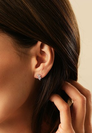 Kvinde rører ved sit mørkebrune hår nær øret, iført små sølvøreringe med tre klare sten mod en beige baggrund.