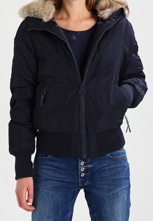 Personne portant une veste noire zippée avec une capuche bordée de fourrure et des poignets côtelés, associée à un jean bleu à boutons.