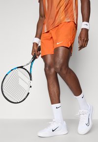 Oranžni atletski kratki hlače z belim Nike logotipom, v kombinaciji z vzorčeno oranžno majico. Belega Nike teniškega čevlja in zapestnic. Črno teniško lopar.