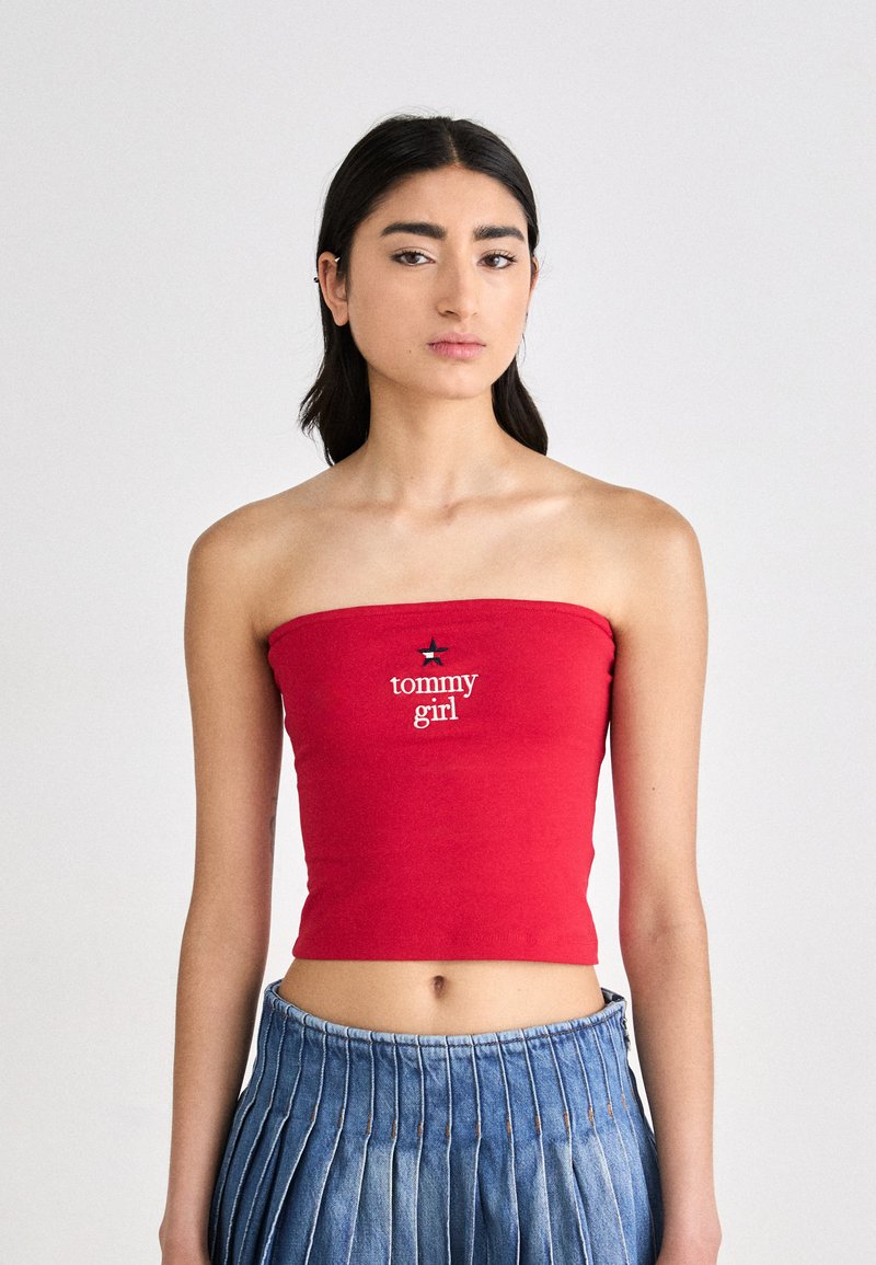 Piros ujjatlan cropped felső, pamutból készült, fehér hímzéssel a "tommy girl" felirattal és csillag kiegészítővel. A modellen pliszírozott kék farmer szoknya van.