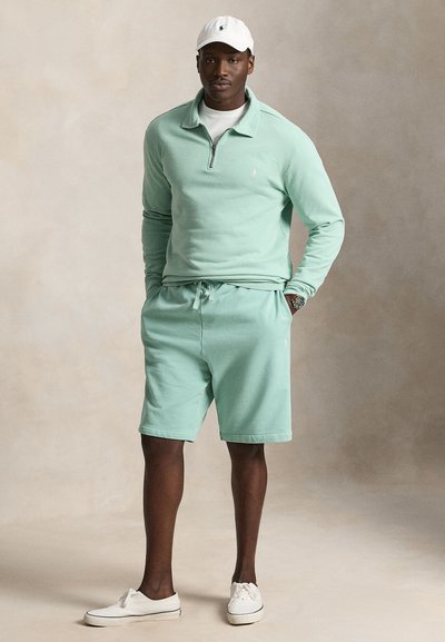 Άνδρας που στέκεται με τα χέρια στις τσέπες, φοράει μιντάτ πράσινο φούτερ με φερμουάρ μέχρι το στήθος, ταιριαστά σορτς, λευκά αθλητικά παπούτσια και λευκό καπέλο του μπέιζμπολ.