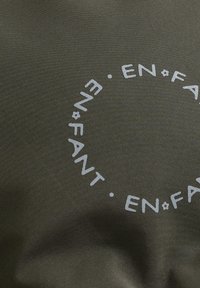 In primo piano, un tessuto verde oliva con un motivo testuale circolare bianco che recita "EN FANT" ripetutamente attorno a piccoli punti.